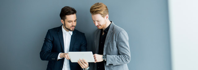 zwei junge Herren im Anzug stehen vor einer grauen Wand und sehen auf ein Tablet